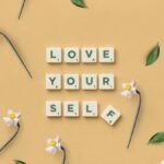 Scrabble tiles spelling 'Love Yourself' surrounded by flowers and leaves on a flat surface.
