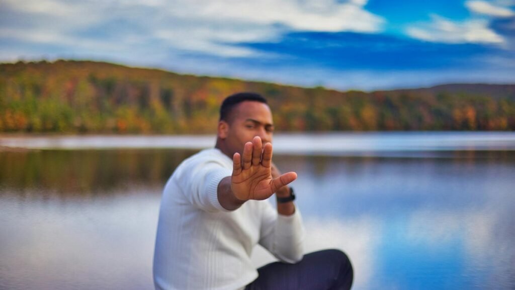 Person sitting by a lake, hand raised in a stop gesture, with autumn foliage in the background.