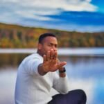 Person sitting by a lake, hand raised in a stop gesture, with autumn foliage in the background.