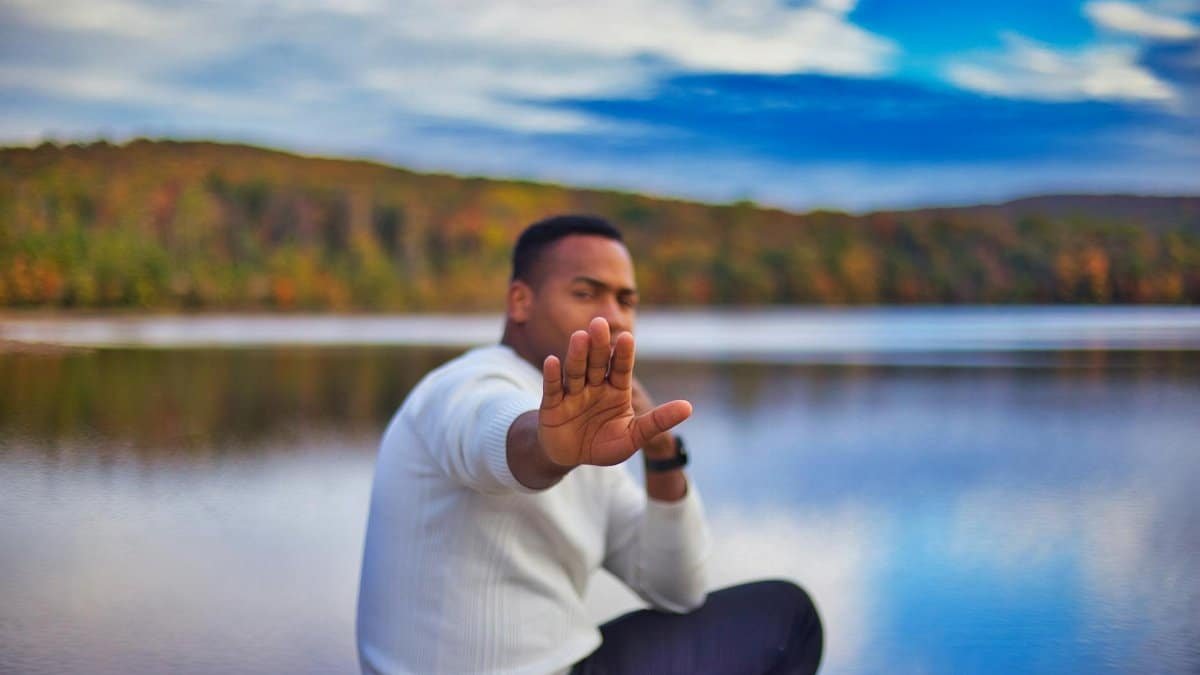 Person sitting by a lake, hand raised in a stop gesture, with autumn foliage in the background.