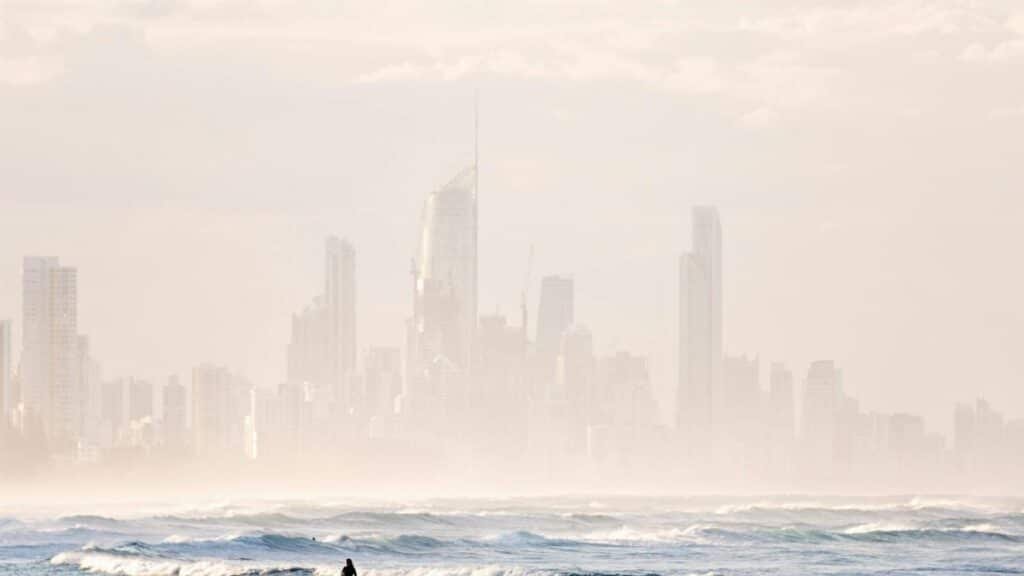 A lone surfer walks along a misty beach with skyscrapers looming in the background, capturing serene coastal urban life.