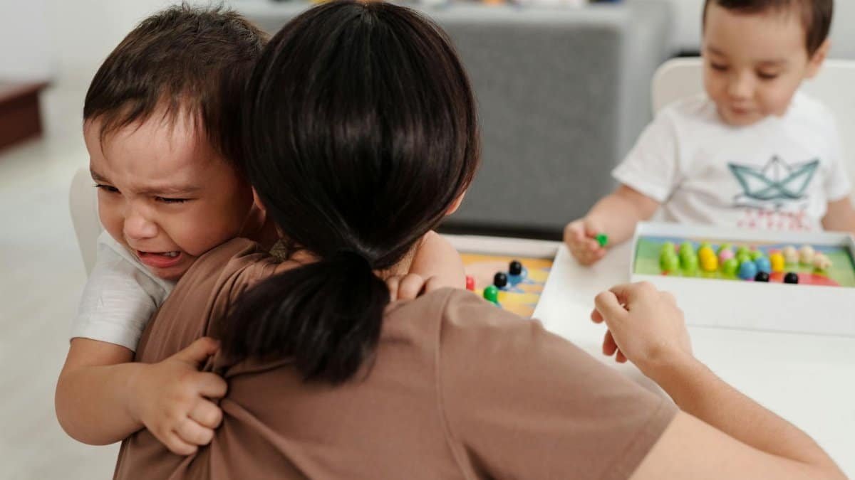 A mother comforts her crying child while another child plays nearby in a cozy home setting.