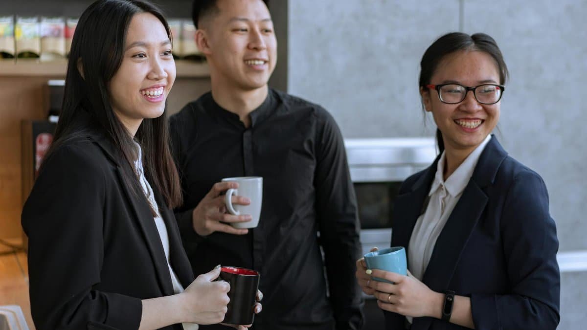 Three Asian coworkers enjoying a relaxed coffee break indoors, smiling and holding mugs.