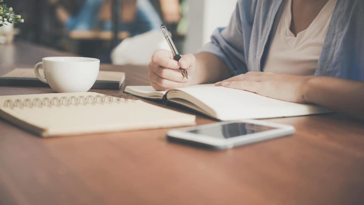 A woman writes in a notebook at a café table with a coffee and smartphone nearby.