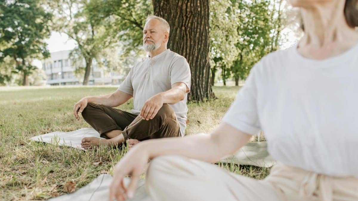 Elderly man and woman meditating outdoors on yoga mats in a peaceful park setting.