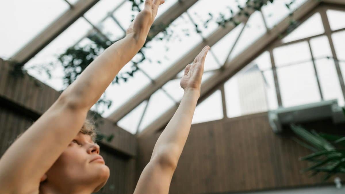 Person practicing yoga indoors with arms raised in a glass-roofed room.