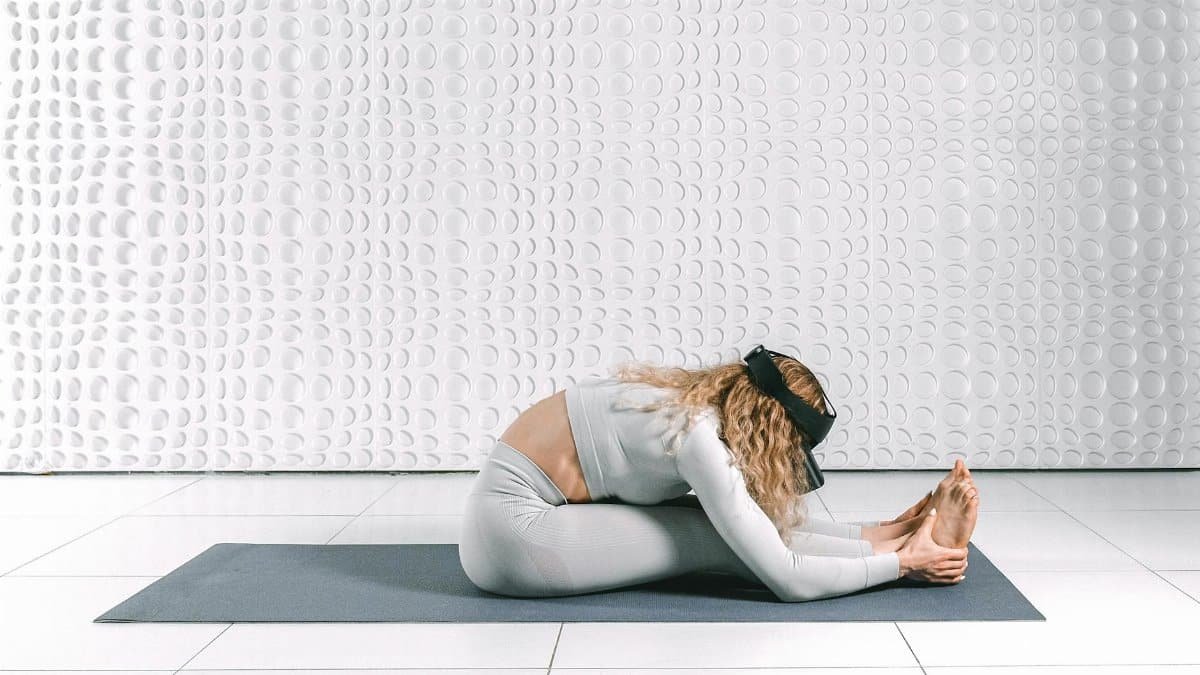 A woman engaging in a yoga stretch while wearing a VR headset in a modern indoor setting.