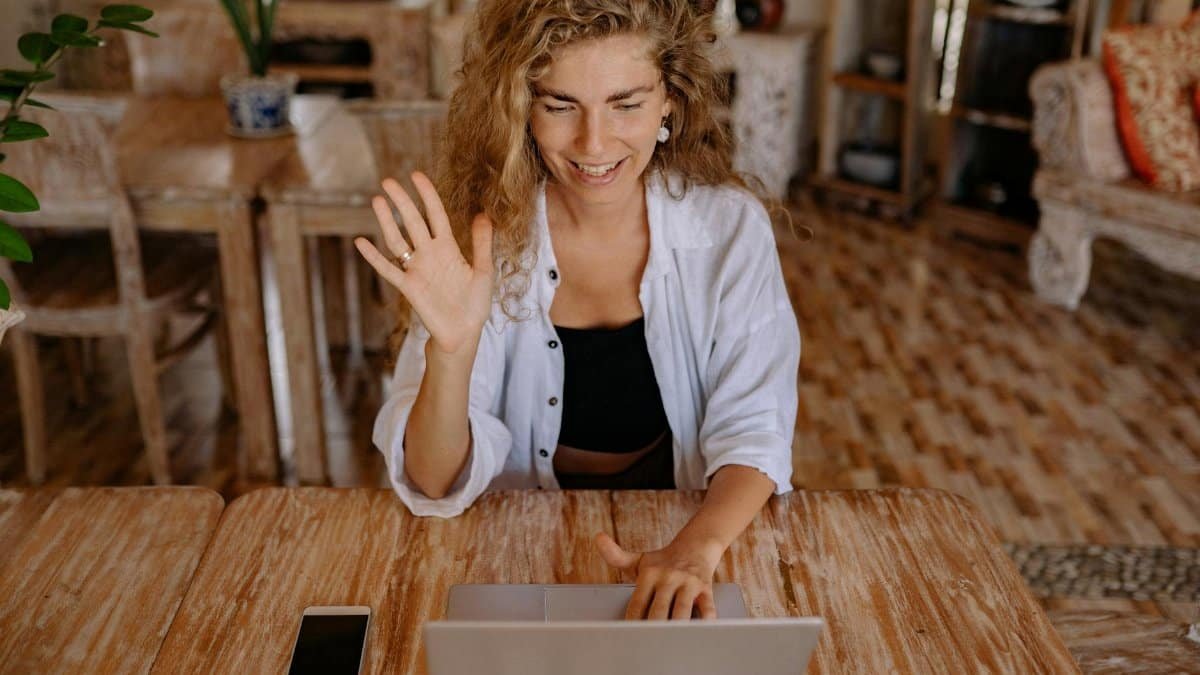 Smiling woman waves during a video call on her laptop in a comfortable indoor setting.