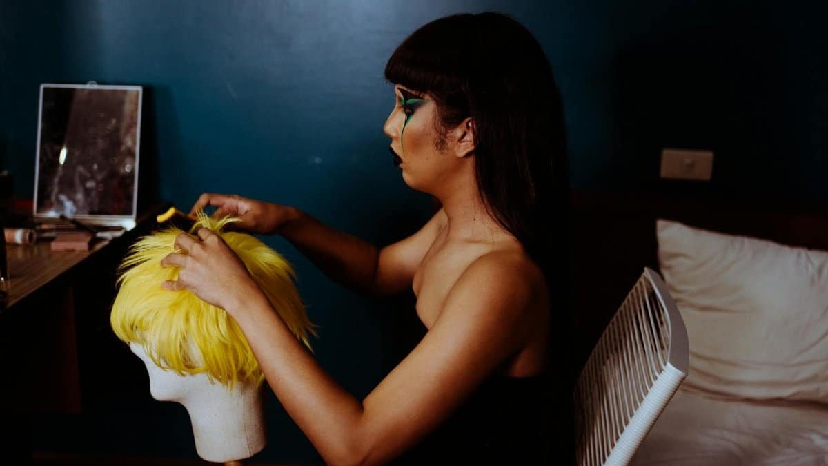 Side view of young male artist with long straight hair and makeup sitting in dressing room and preparing hairpiece for performance