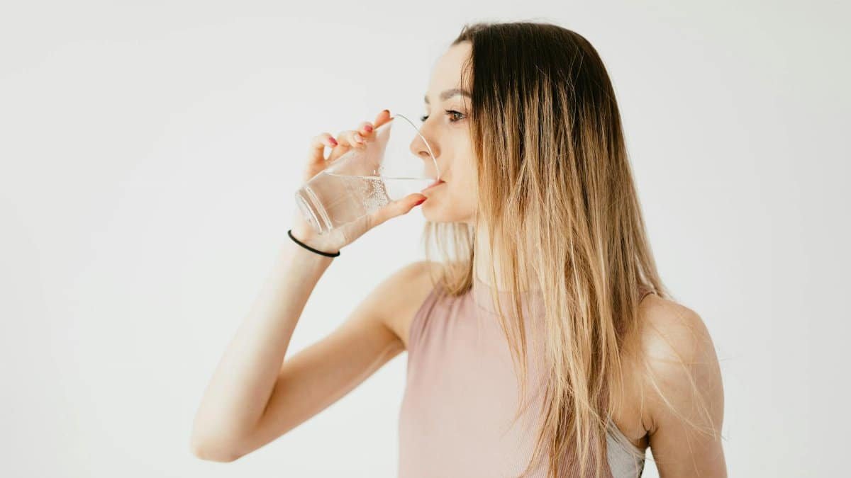Young thirsty fit female with long hair in sportswear drinking water while recreating after workout
