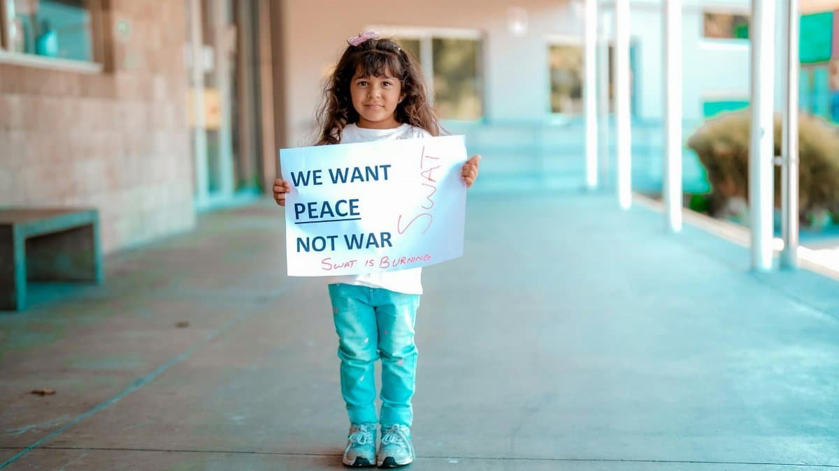 Little girl with long hair standing on pavement holding a 'We Want Peace Not War' sign.