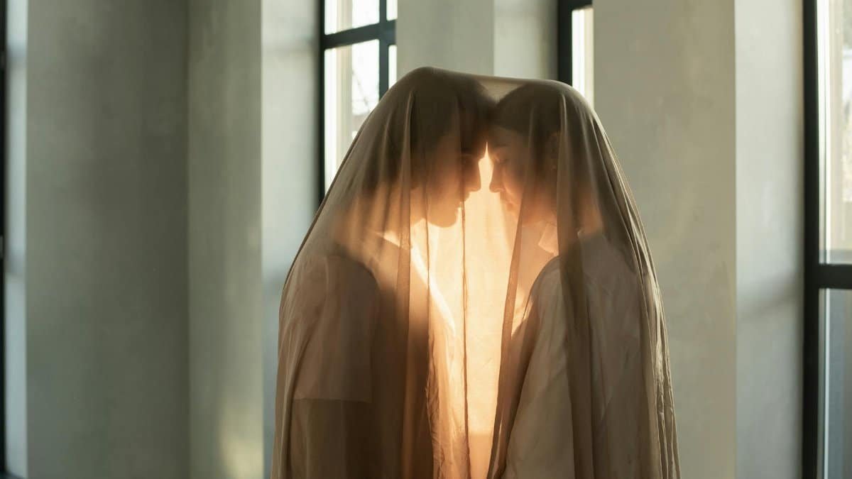 Two people in a romantic embrace under a sheer veil indoors, bathed in warm light.