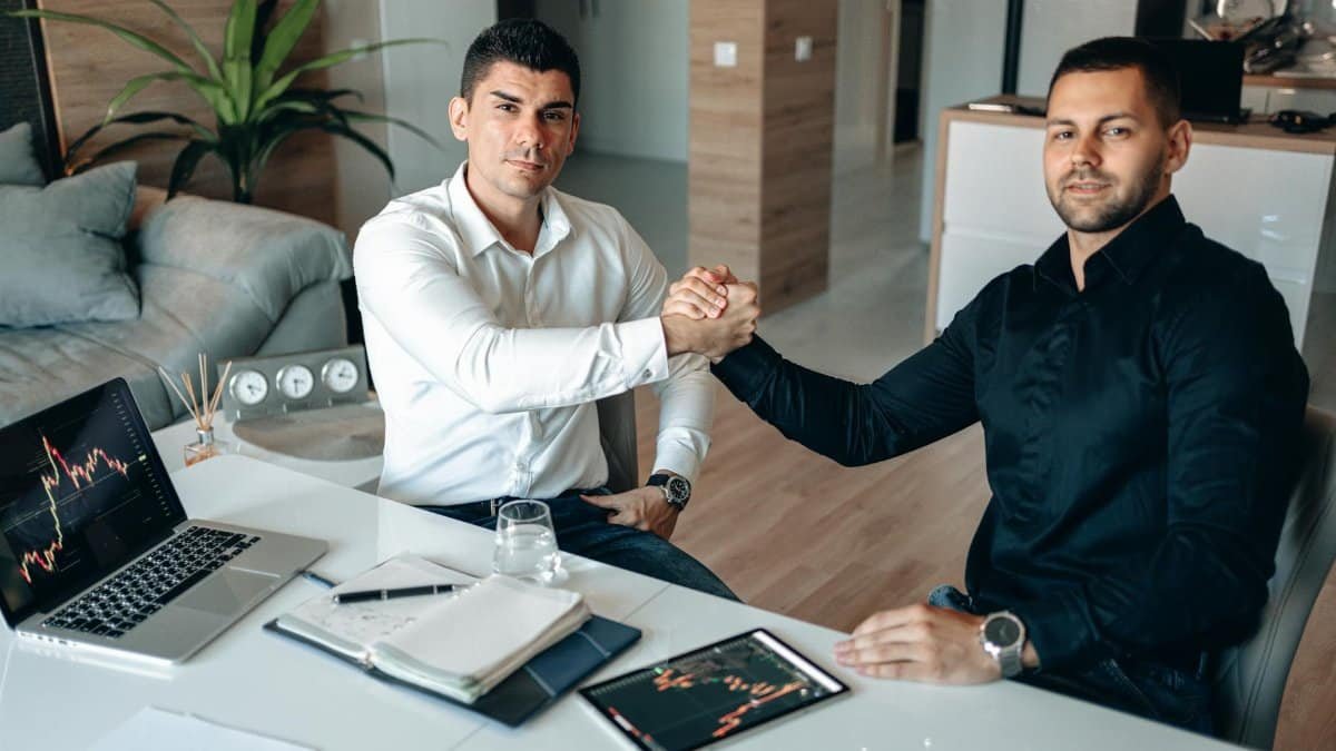 Two businessmen shaking hands at a modern office desk with tech devices, indicating a successful partnership.