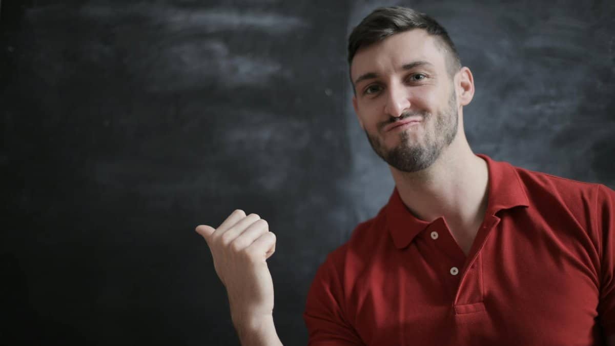 Portrait of a young man in a red polo, gesturing confidently indoors with a chalkboard background.