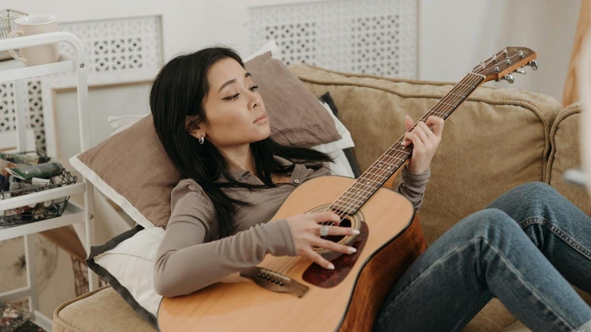 Asian woman lounging on a sofa playing an acoustic guitar. Relaxing home music session.