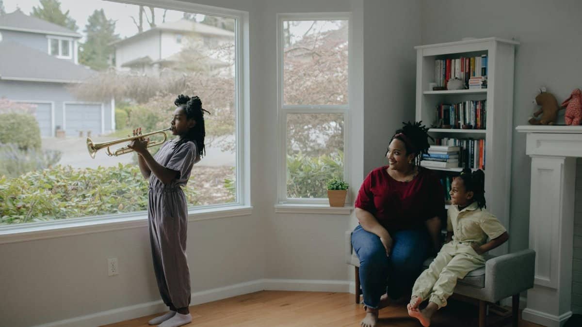 Child playing trumpet indoors as mother and sibling watch, capturing a family moment with music.