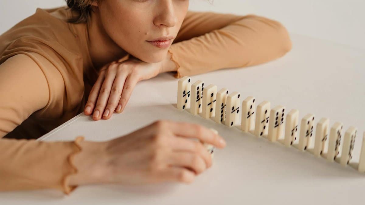 A woman in focus, contemplating while playing with dominoes on a white table.