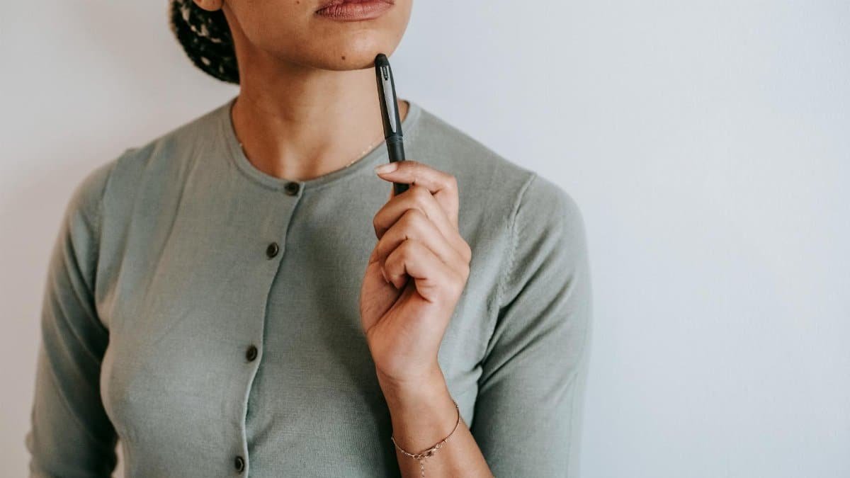 Crop anonymous ethnic female with dark hair in casual outfit touching face with pen and thoughtfully looking away against white wall