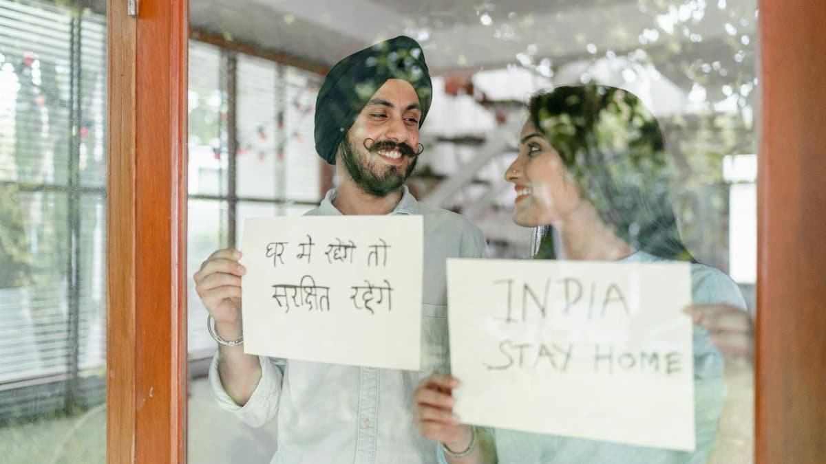 Through glass of young Indian couple standing against window and looking at each other while demonstrating sheets of paper with India stay home inscription in English and Hindi