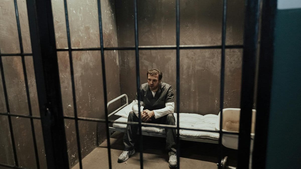 A man sits alone on a bed in a dimly lit prison cell, looking contemplative and isolated.