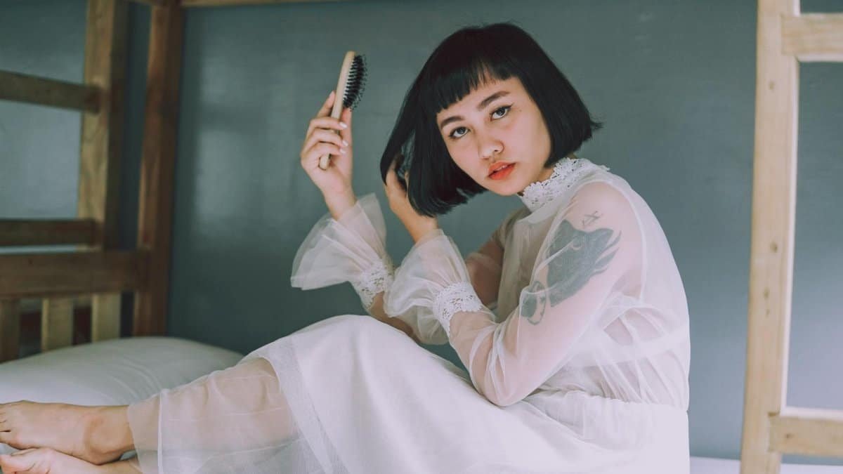 Stylish woman with bob haircut poses in sheer white dress, holding a hairbrush in a relaxed indoor setting.