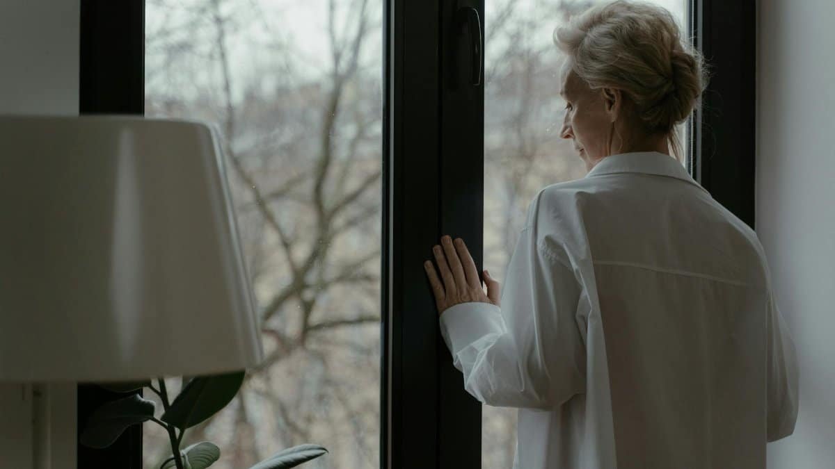 A senior woman in white looking out a window thoughtfully, symbolizing reflection.