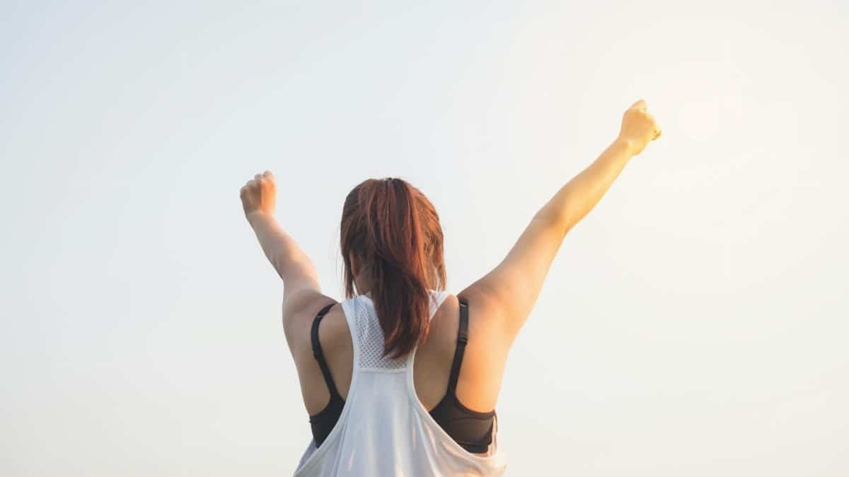 A woman celebrates success with arms raised in a bright outdoor setting.