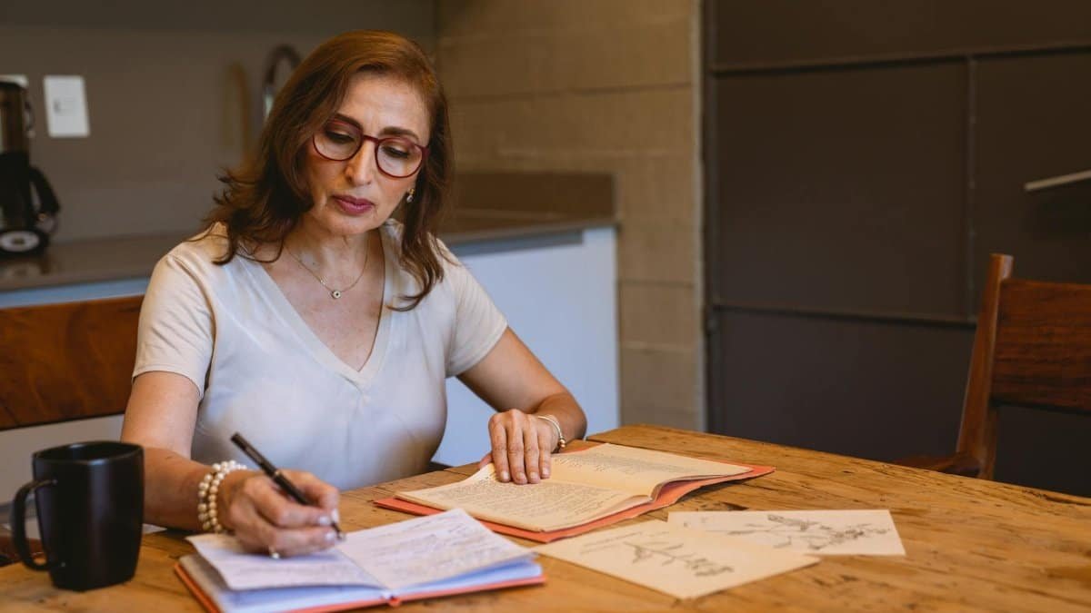 Adult woman writing notes in a cozy kitchen setting with books and a cup of coffee.