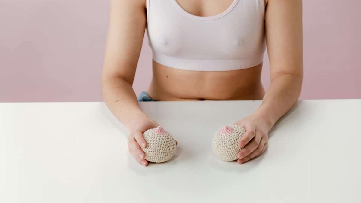 A woman in a sports bra holds knitted breast models on a pink background, symbolizing breast health awareness.