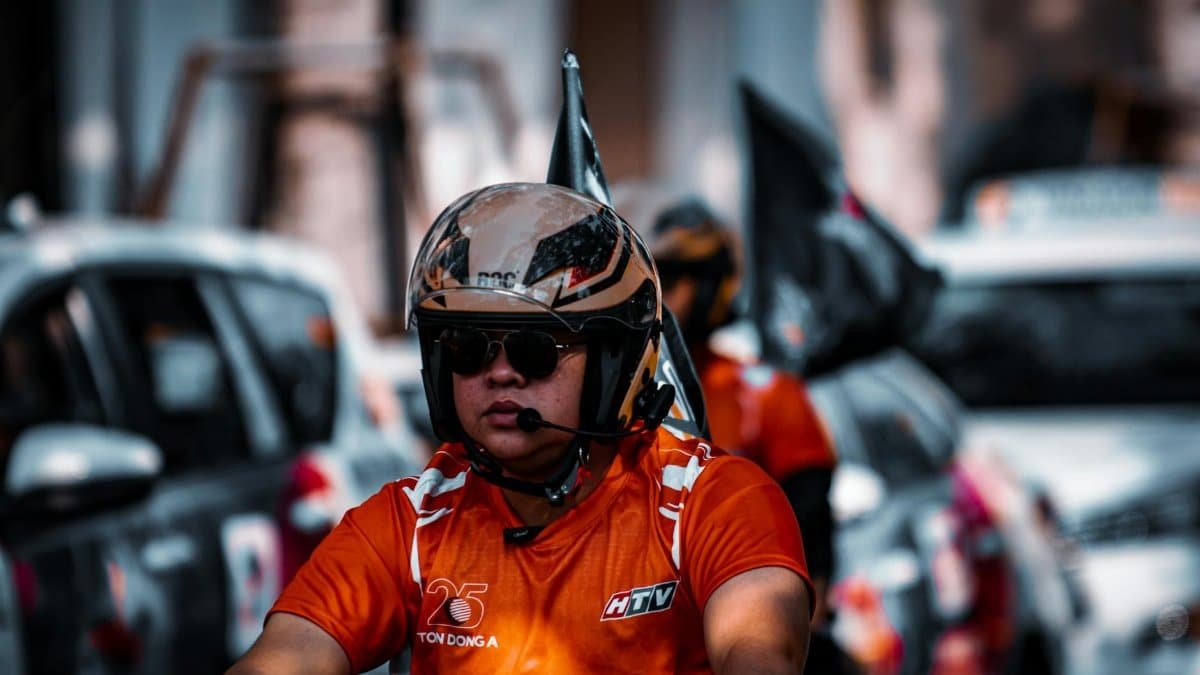 Motorcyclist in orange jersey and helmet navigating city traffic, exhibiting urban culture and transport.