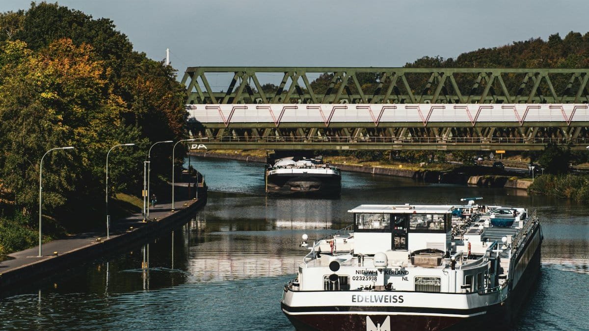 Large river barge navigating a scenic European canal beneath an iron bridge.