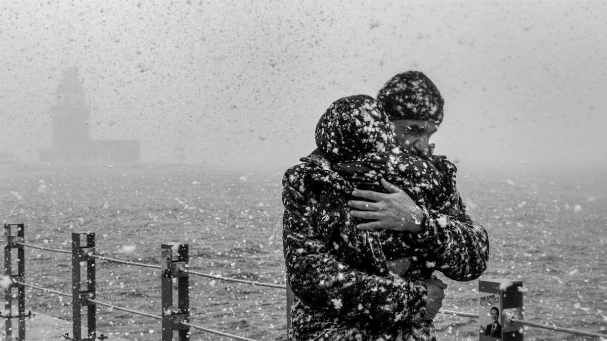A moving black and white photo of a heartfelt embrace during a snowfall at the waterfront.