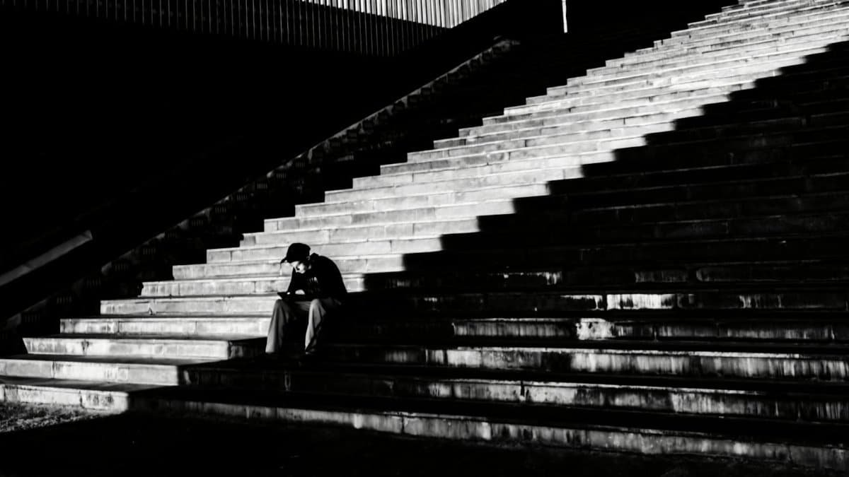 A solitary person sits pensively on urban steps at night, creating a dramatic black and white composition.