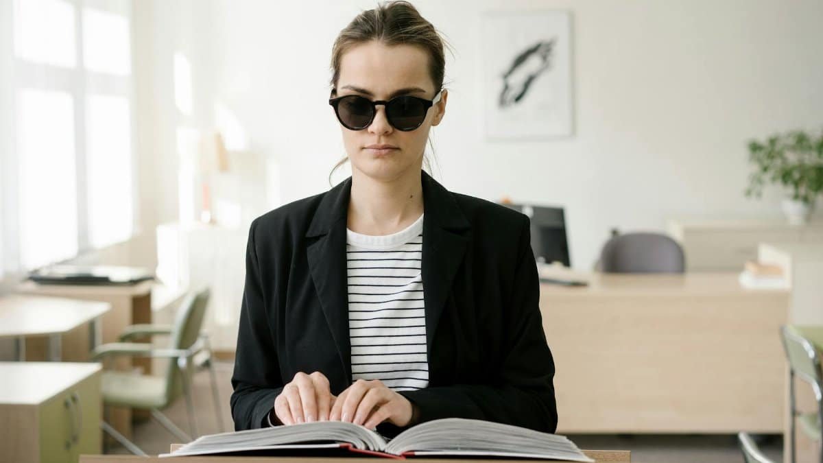Blind woman wearing sunglasses reads a braille book at a podium in an office.