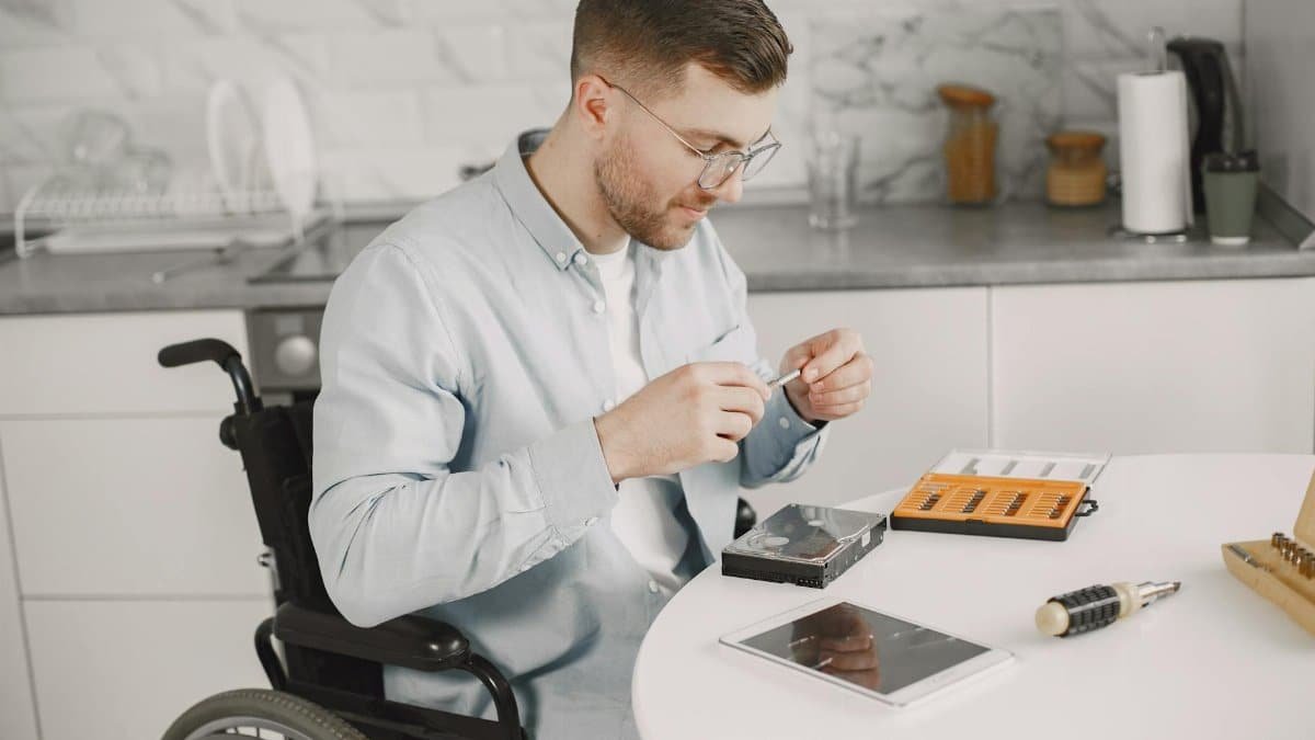 Man in wheelchair repairing electronics at home using tools, showcasing independence.