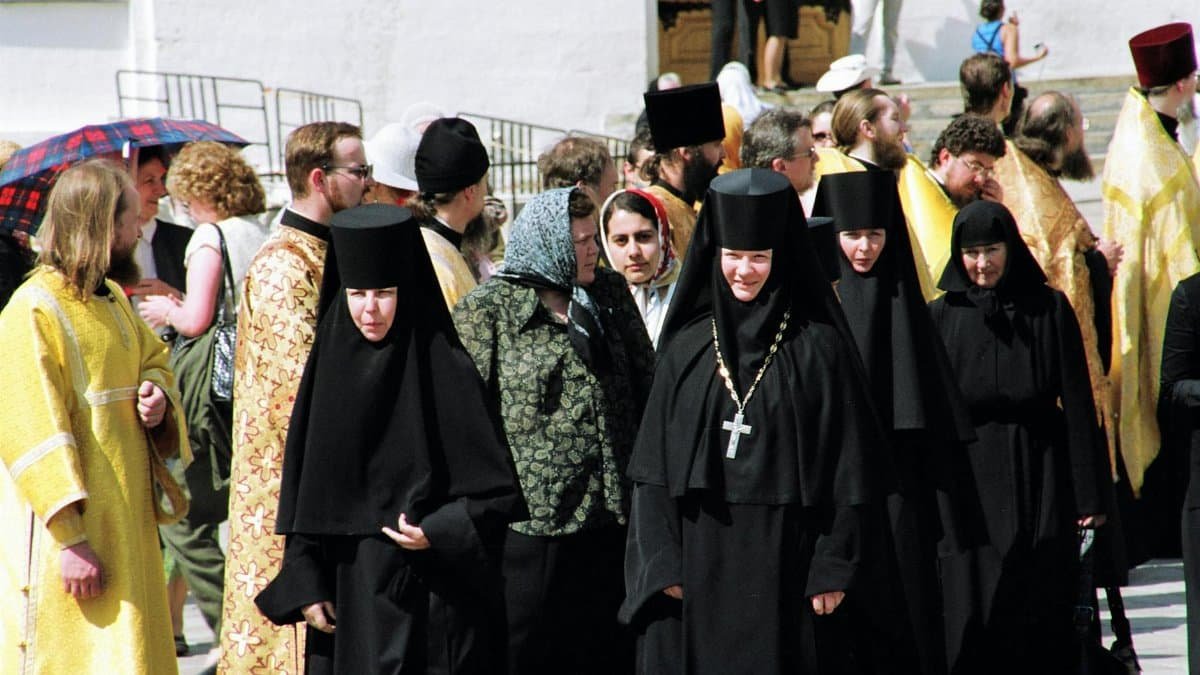 Nuns of convent wearing black clothes walking along street passing men of cloth in golden color robes and common people located near building celebrating holiday in sunny day