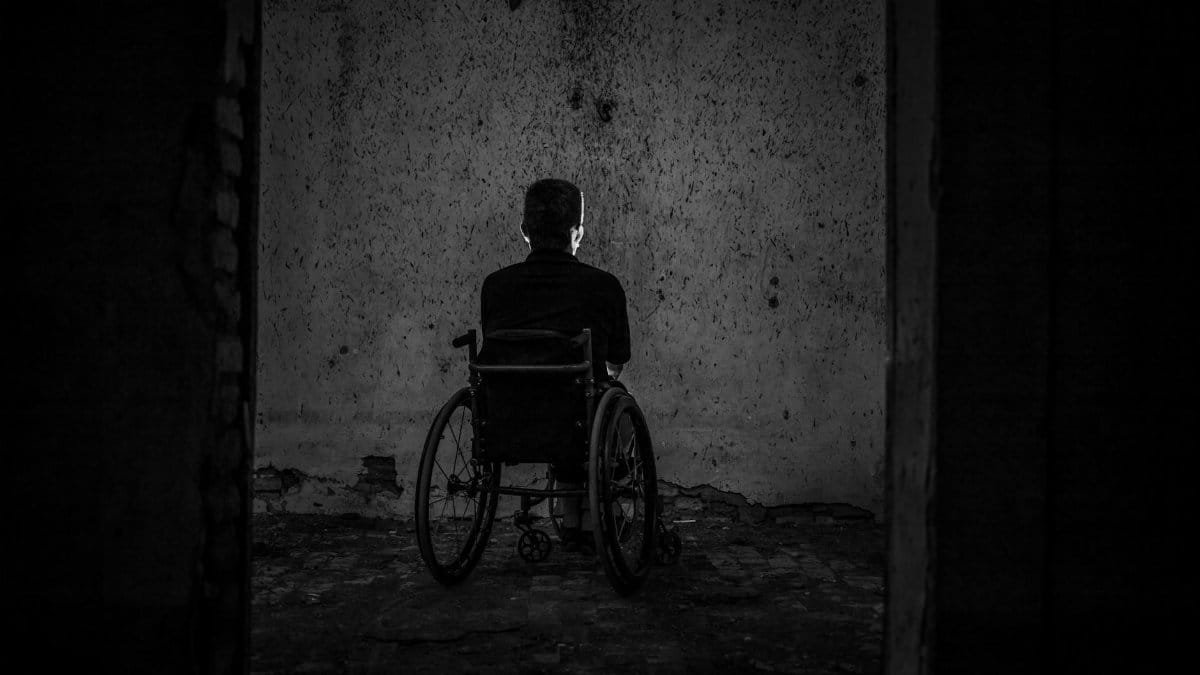A man sits alone in a wheelchair facing a wall in a dark, empty room, evoking solitude.