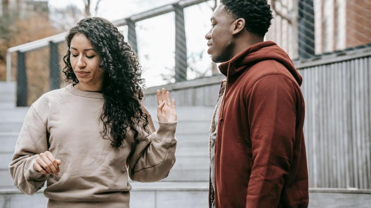Upset ethnic girlfriend with raised hand and concerned African American boyfriend breaking up on street against metal grid on background