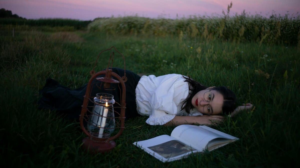 Woman relaxing with a book and lantern in a tranquil field at twilight, offering a serene escape.