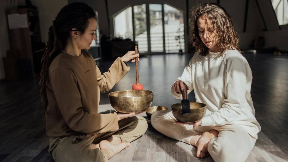 Two women sitting indoors meditating with singing bowls, promoting relaxation and healing.