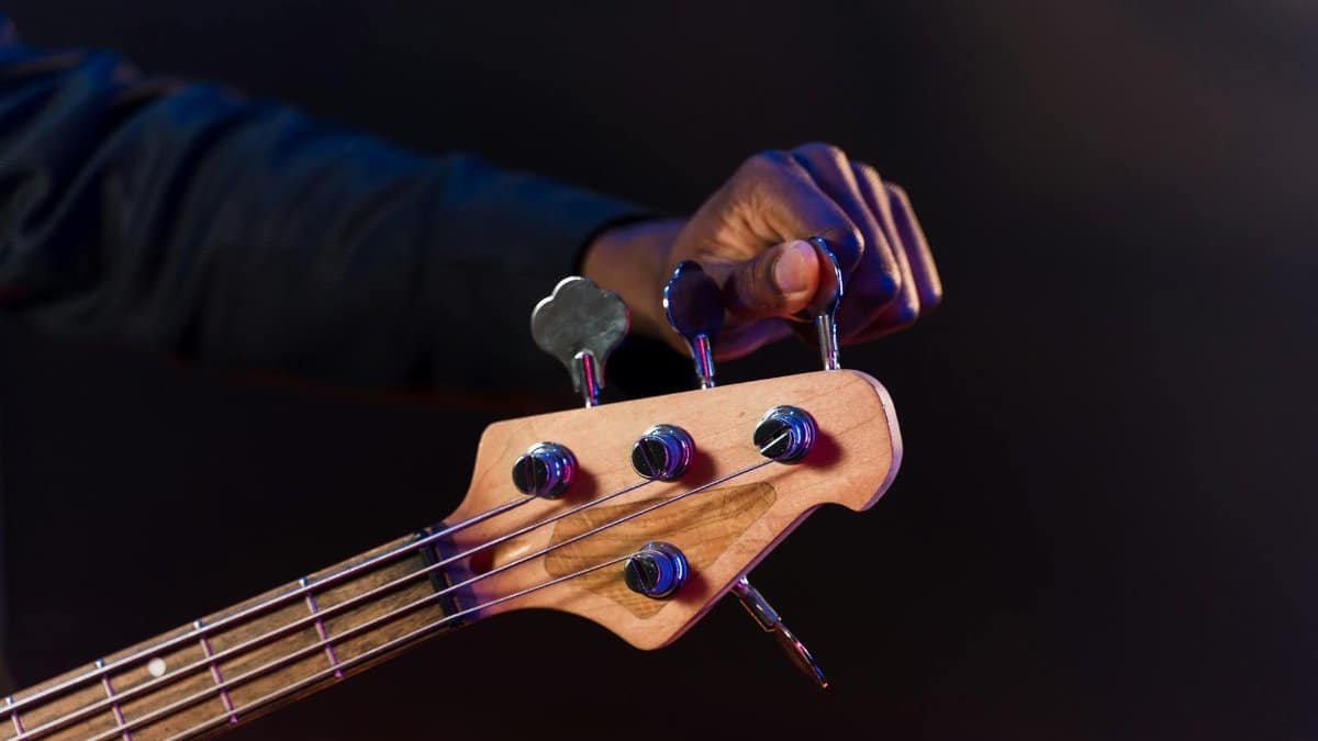 Detailed shot of a hand adjusting tuning pegs on a bass guitar's headstock with a dark background.