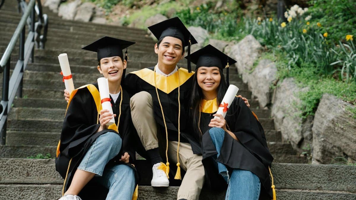 Three happy graduates in caps and gowns celebrate on outdoor steps, holding diplomas.