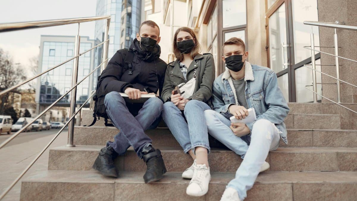 Three young adults seated on urban steps, wearing masks for protection.