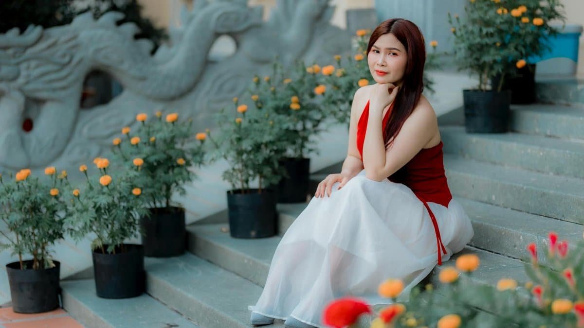 Elegant woman in a red dress sitting amidst vibrant flowers on stone steps outdoors.