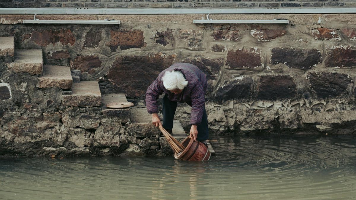 An elderly person gathering water from a pond beside a rustic stone wall and stairs.
