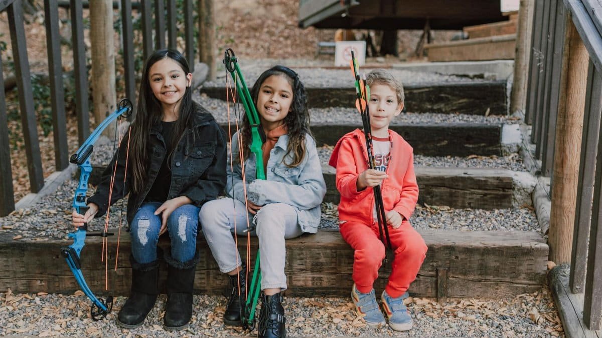 Three children with bows and arrows practicing archery outdoors, smiling and sitting on wooden steps.