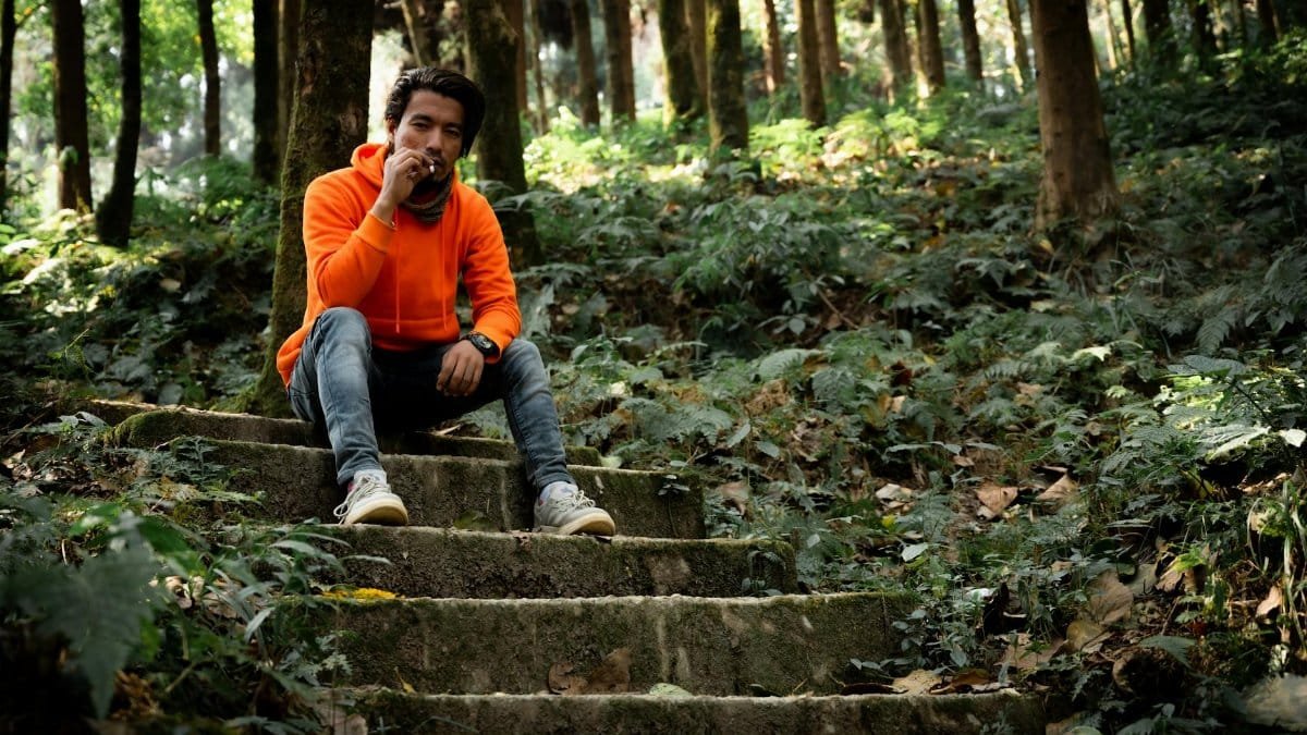 Man in an orange hoodie sits smoking on forest steps in Tukdah, India, surrounded by lush greenery.