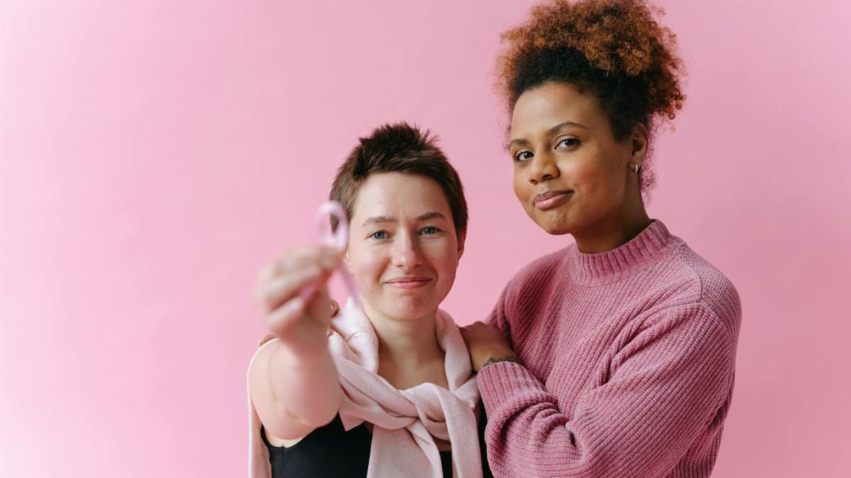 Two women embracing, one holding a pink ribbon for breast cancer awareness, against a pink background.