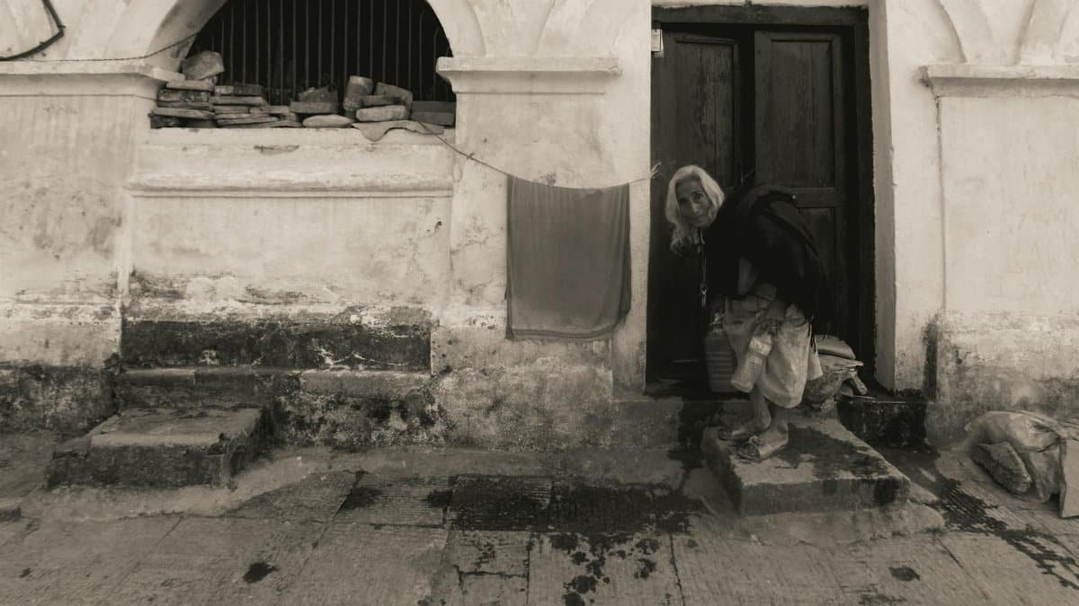 A grayscale image of an elderly woman standing at a weathered doorway of a historic building.
