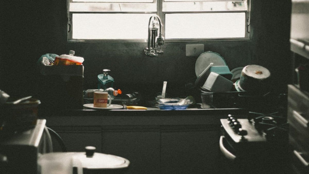 A cluttered kitchen sink filled with dirty dishes and utensils near a window, creating a messy home interior scene.